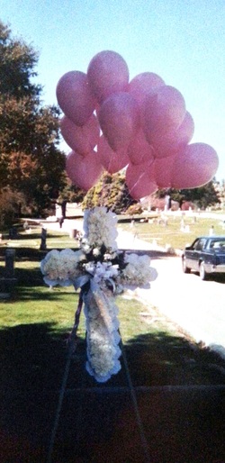 White flower cross at grave site with ppink balloons at baby's funeral